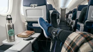 A traveler on an airplane with their legs stretched out, wearing blue sneakers and heated socks, with a portable battery on the tray table.